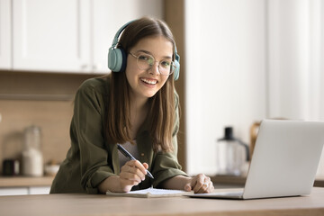 Portrait of cheerful pretty teenager school student girl wearing earphones and eyeglasses, working...