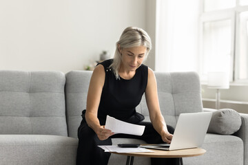 Serious blonde mature accountant woman reviewing paper bills, invoices, paying taxes, fees,...