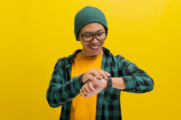 Excited Asian man in a beanie hat, casual shirt, and eyeglasses celebrates success on time while checking his wristwatch. Isolated on a yellow background