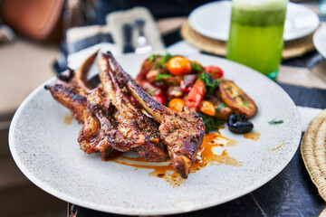 Gourmet lamb chops on a white plate with roasted vegetables, asparagus, and a green drink on a...