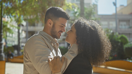 A man and woman embrace lovingly outdoors, amid city greenery under sunlight.