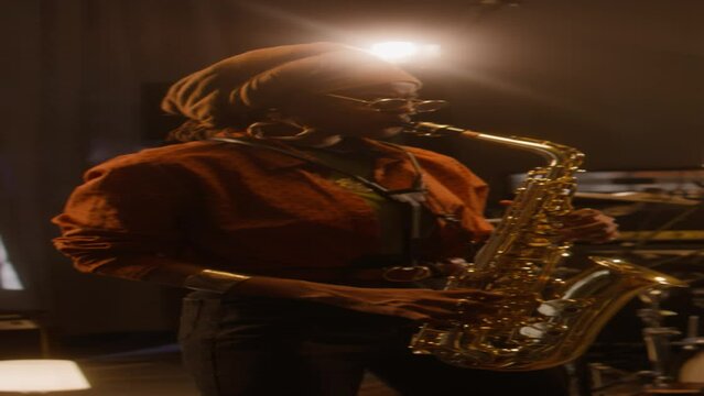 Medium vertical of African American female jazz band member in ethnic turban and glasses playing saxophone solo, while performing at concert or rehearsing at recording studio