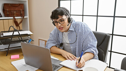 A bespectacled man with a headset works intently at a laptop in a contemporary office, writing...