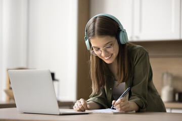 Happy teen high school student girl in glasses and earphones studying at laptop computer, writing...