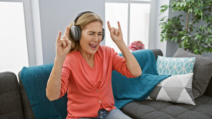 A jubilant mature woman enjoys music on headphones in a cozy living room interior, expressing joy...