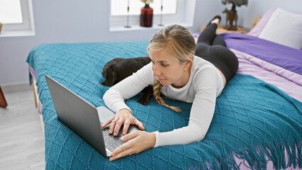 A young woman lying on a bed with her labrador dog beside her, working on a laptop in a cozy...