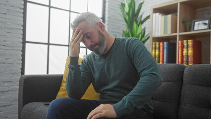 Stressed man sitting in modern living room with hand on forehead, expressing anxiety or headache...