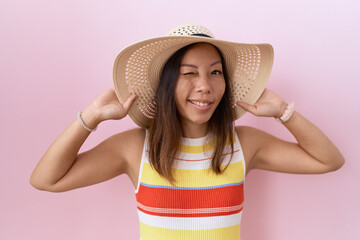 Middle age chinese woman wearing summer hat over pink background winking looking at the camera with...