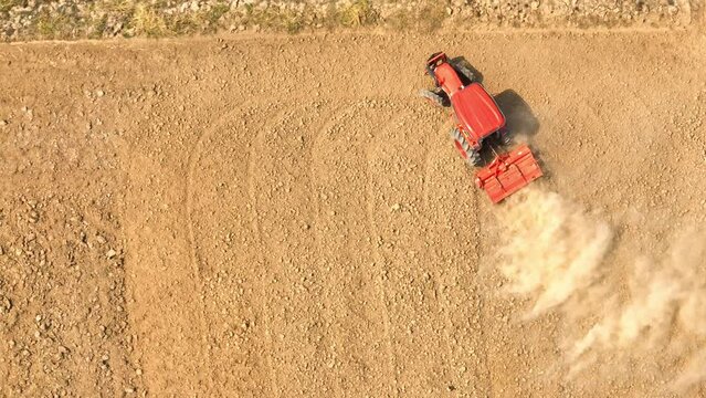 An aerial vista captures a tractor guiding a cultivator across a sprawling, parched landscape, casting up billows of dust that underscore the earth's desiccated texture against a flawless sky.
