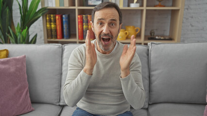 A surprised middle-aged man with a beard sitting on a sofa in a cozy living room expressing...