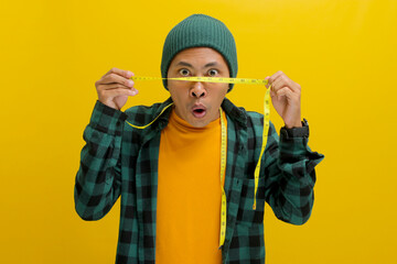 An enthusiastic young Asian man, clad in a beanie hat and casual shirt, gazes at a measuring tape with an expression of surprise or excitement, mouth slightly agape, against a yellow background