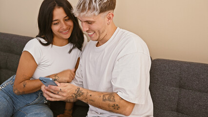 A smiling man and woman enjoy time together on a couch, looking at a smartphone in a cozy living...