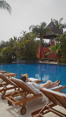 A young woman relaxes on a lounger by a resort's blue pool in bali, surrounded by tropical foliage.