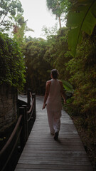A woman walks on a wooden pathway surrounded by lush tropical vegetation, epitomizing serene resort...