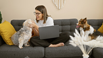 Hispanic woman working on laptop with two dogs in a cozy living room, depicting a comfortable...