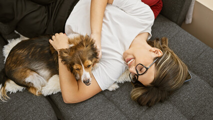 A young hispanic woman smiling while cuddling with her dog on a couch inside a cozy living room.