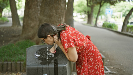 Gorgeous hispanic woman quenching thirst with refreshing drink from park fountain, her glasses...