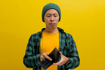 Frowning young Indonesian man holds an empty wallet, isolated on a yellow background