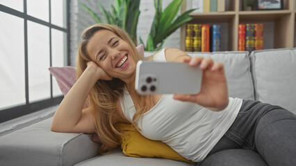 A cheerful young woman takes a selfie on a cozy sofa in a stylishly decorated living room, exuding...