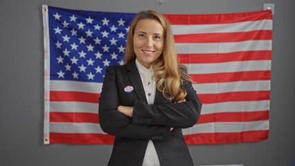 Smiling blonde woman with crossed arms wearing a 'voted' sticker, standing before an american flag in an office.