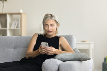 Relaxed blonde middle aged woman with cellphone resting on home couch, texting message on smartphone, chatting, typing, using online application Internet technology