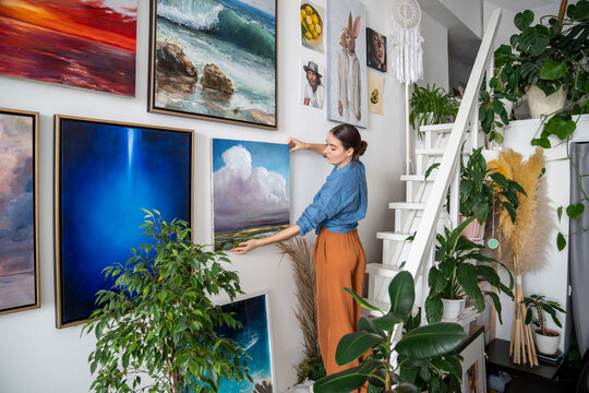 Serious woman hangs pictures on wall. Concentrated unsmiling female artist creates composition of paintings, decorations on wall. Creative designer in art home studio workshop among artworks plants.