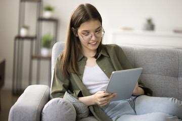 Positive smart teenager schoolkid girl in glasses using modern technology, educational app on tablet, playing online game, watching Internet content, browsing social media