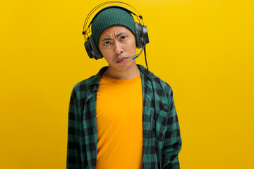 Asian man in a beanie and casual clothes, deep in thought with pensive expression while listening to music on headphones. Isolated on a yellow background.