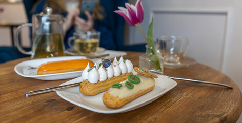 A casual café setting with a wooden table featuring an eclair on a white plate, accompanied by silver cutlery and a glass cup.