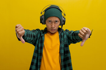 Asian man in a beanie and casual clothes frowns in disapproval while listening to music on his...