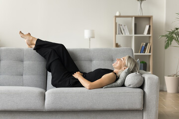 Positive serene mature woman lying on couch with closed eyes and happy smile, placing legs and feet...