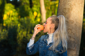 Pretty young blonde woman with a ponytail in her hair wearing a denim dress holds an orange in her...