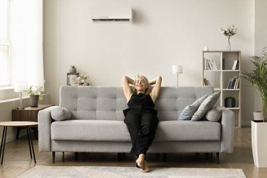 Positive homeowner woman enjoying leisure, ventilation, comfortable cool air temperature under conditioning split system at home, resting with closed eyes on soft stylish couch,
