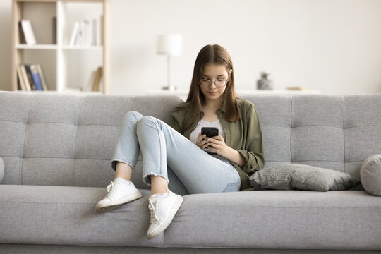 Focused gen Z teenage schoolkid girl in glasses using online educational application on smartphone, relaxing on sofa, enjoying Internet technology, leisure with digital gadget at home
