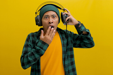 Asian man in a beanie and casual clothes, wearing headphones, looks shocked with his mouth agape...