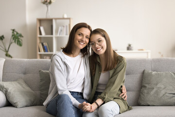Obraz premium Happy pretty teenage kid girl and beautiful mom sitting together on sofa, looking at camera, smiling. Mother holding daughters hand, hugging teen child with care, love. Home family portrait