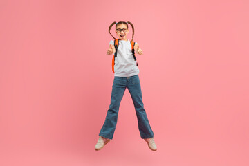 A young girl is energetically jumping in the air with a backpack securely strapped on her back, showing thumb ups, pink studio background