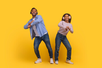 African American man and woman are captured mid-dance, both smiling and enjoying themselves as they perform a coordinated movement, using headphones on yellow background