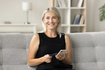 Happy pretty blonde mature woman holding credit card and mobile phone, looking at camera at home, smiling for bank customer portrait, promoting financial online service, cashback, easy payment