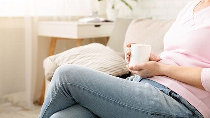 A close-up of woman sitting comfortably on a beige sofa, casually holding a white mug in her hand,...