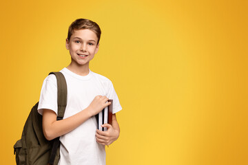 A young boy standing while holding a book and a backpack.