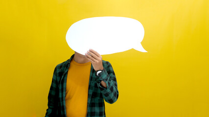 Asian man, concealing his face behind a speech bubble held in front of him, stands against a yellow background, depicting the concepts of information, thoughts, quotes, opinions, reviews, and feedback © Jamaludinyusup