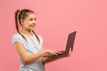 A young girl stands in front of a pink background, holding a laptop computer in her hands. She...