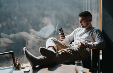 A calm adult male leisurely sits in a chair, enjoying the morning sun while using a smart phone in...