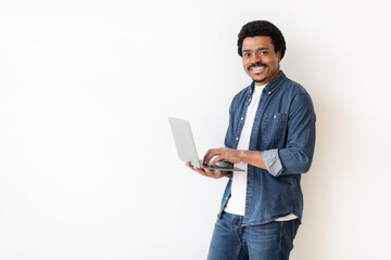 African American man is standing, holding a laptop in his hands. He is looking at the screen intently, possibly working or browsing. The background is white with copy space