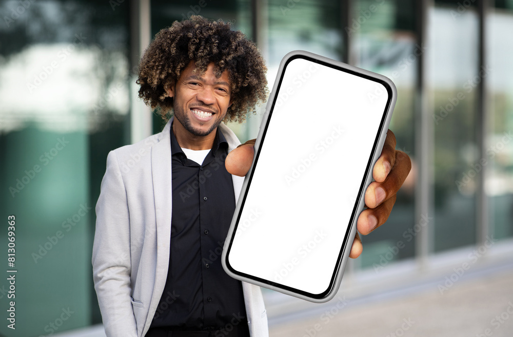 Poster African American man is standing while holding a cell phone in his hand, looking at the screen. He appears focused and engaged with the device, possibly texting or browsing.