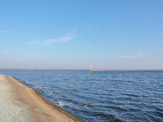 Old concrete lighthouse on the edge of the Kyiv sea spit, calm blue water with distant white yacht and blue sky with feather clouds, narrow bank strip on horizon.