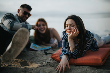 Three friends having fun on a sandy beach, one woman lies down while a couple sits beside her,...