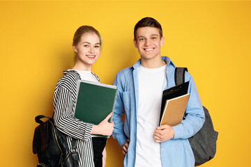 Two happy teenagers are standing side by side against a vibrant yellow backdrop, carrying backpacks and holding books