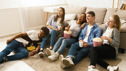 Several friends sitting on the floor together, each holding a bag of popcorn and eating. The group...